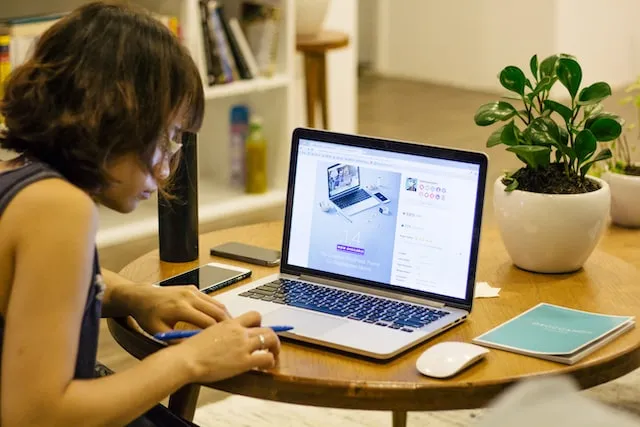 Kobieta siedząca przy biurku i patrząca na laptopa stojącego przed rośliną domową w białej doniczce