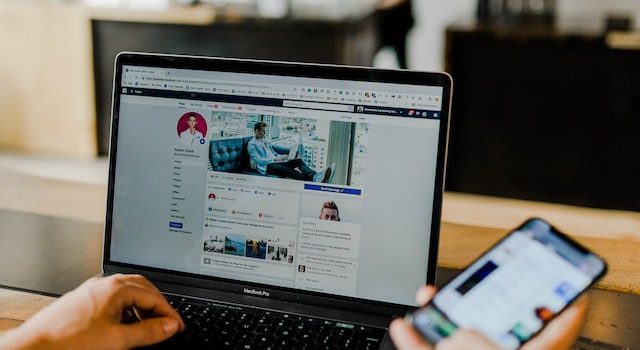 Bir elinde akıllı telefon tutan ve sol elini kullanarak bir dizüstü bilgisayarda Facebook'ta gezinen kişi
