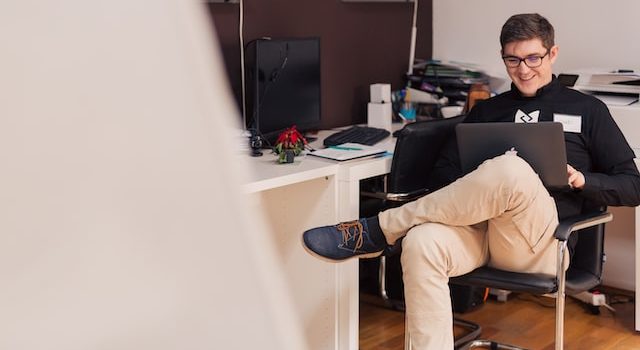Mężczyzna siedzący na krześle obok biurka i patrzący na laptopa, który trzyma na kolanach