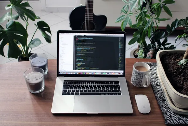 Un laptop sulla scrivania con una tazza di caffè, due candele e un mouse bianco accanto