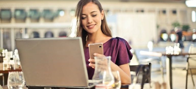 Kobieta siedzi w kawiarni, pracuje na laptopie i trzyma smartfon w lewej ręce