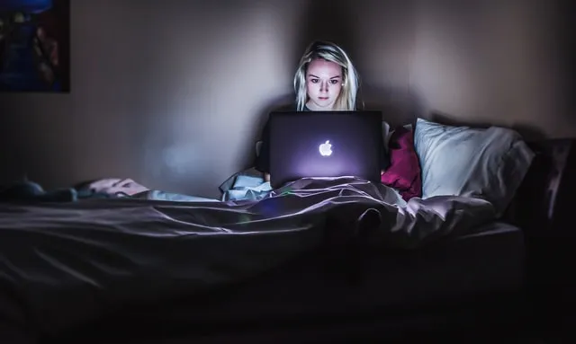 Uma mulher loira deitada na cama e olhando para seu MacBook no escuro
